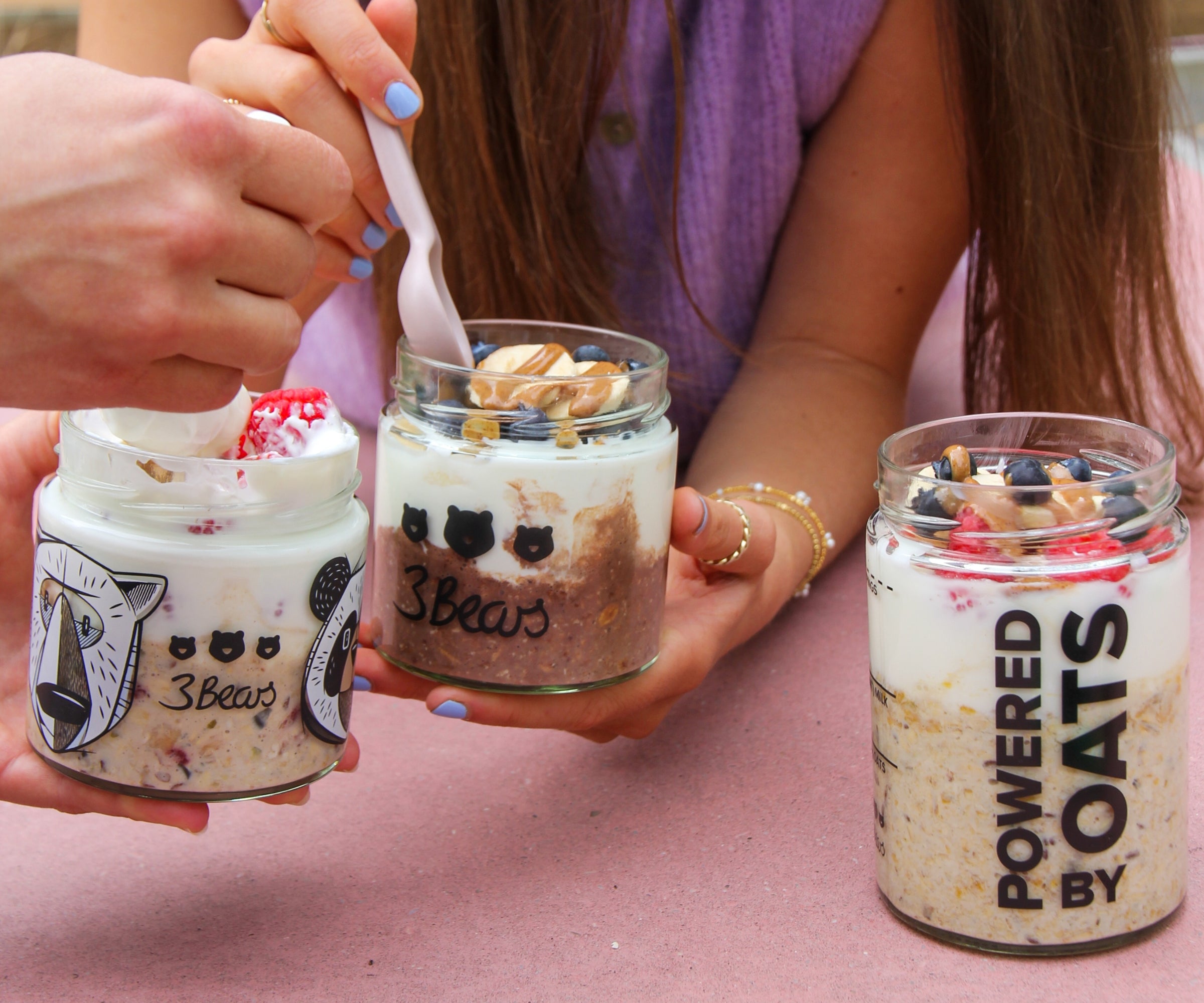 People holding three jars with different Overnight Oats flavours, lying on a picnic bench